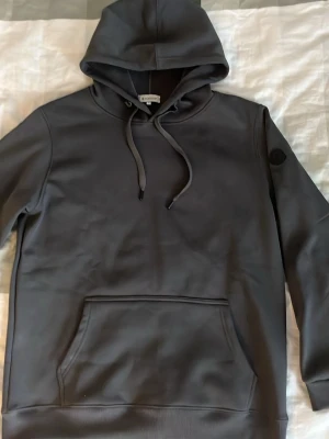 Svart/grå hoodie från Moncler - Snygg svart hoodie från Moncler med klassisk huva och dragsko. Hoodien har en stor magficka och en diskret Moncler-logga på ärmen. Tillverkad i mjukt material som känns skönt mot huden. Perfekt för dig som gillar stilrena och exklusiva plagg. Har däremot lite fläckar sen innan på ryggen 