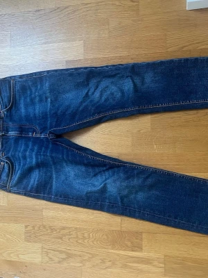 Slim fit jeans från Nudie Jeans - Säljer ett par mörkblå jeans från Nudie Jeans med klassisk femficksdesign och snygga slitningar. Perfekt för dig som gillar en tidlös och cool look. Storlek 30W/32L och modellen är Lean Dean