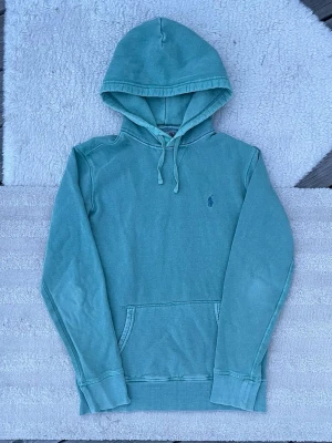 Turkos hoodie från Polo Ralph Lauren - Grön hoodie från Polo Ralph Lauren med klassisk logga broderad på bröstet. Hoodien har en stor huva med dragsko och en praktisk magficka. Materialet är mjukt och känns som bomull, perfekt för chill dagar. Har lite små slitningar men bidrar till en vintage vibe