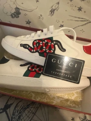 Gucci Ace sneakers med orm - Säljer ett par vita Gucci Ace sneakers i läder med den ikoniska broderade ormen på sidan och gröna/röda ränder. Skorna har vita snören, röd och grön detalj på hälen och klassisk låg profil. Perfekta för dig som vill sticka ut med lyxig streetstyle.
