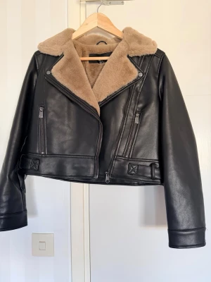 Svart skinnjacka med beige foder - Cool svart skinnjacka från Vero Moda med mjukt beige foder och krage. Jackan har flera dragkedjor, sned knäppning och detaljer som spännen vid midjan. Perfekt för dig som vill ha en edgy look med lite extra värme.