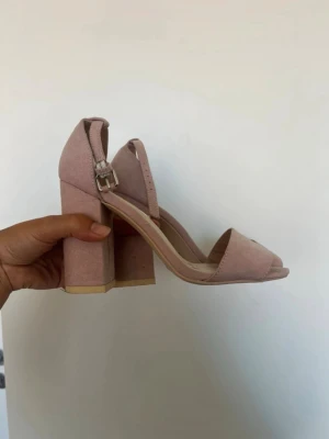 Rosa chunky klackpumps med rem - Säljer ett par ljusrosa pumps med öppen tå och bred klack. Skorna har en justerbar ankelrem med silverfärgat spänne och är tillverkade i mockaimitation. Perfekta för dig som gillar en trendig och bekväm look.