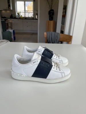 Vita valentino open sneakers med blå detalj - Snygga vita valentino open sneakers från Valentino med bred mörkblå läderdetalj över snörningen och coola nitar på hälen. Skorna har en klassisk låg profil och är tillverkade i läder, vilket ger en lyxig känsla och stilren look. Skorna är i riktigt bra skick och är i storlek 39 men kan passa 40. Skorna är köpa från Vestiaire collective och de är självklart äkta. Vid frågor eller funderingar är det bara att säga till Mvh.
