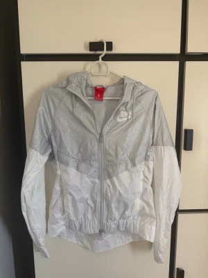 Nike vit och ljusgrå vindjacka - Nike vindjacka i vit och ljusgrå färg med dragkedja och huva. Jackan har meshfoder, elastisk nederkant och logotyp på bröstet. Perfekt för att hålla vinden borta under utomhusaktiviteter. Materialet är lätt och vattenavvisande.