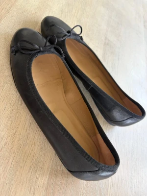 Svarta ballerinaskor från Marzio - Stilrena svarta ballerinaskor från Marzio med rund tå och dekorativ rosett framtill. Skorna har en slät yta i syntetmaterial och en mjuk innersula i beige. Perfekta för dig som gillar klassisk och enkel design.