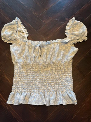 Blå blommig topp med puffärmar - Söt ljusblå topp med små vita blommor, puffiga ärmar och volangdetaljer. Smockad midja och volangkant nedtill, samt knytband vid halsen. Perfekt för en romantisk och trendig look.