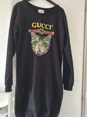 Svart långärmad klänning med paljetter för barn - Cool svart långärmad klänning från med ett stort glittrigt kattmotiv och gröna ögon på bröstet. Klänningen har logotypen och klassiska färger ovanför motivet. Materialet är mjukt och bekvämt, och så himla gulligt att ha på sig för ditt barn. :)
