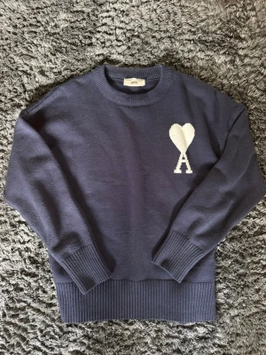 Mörkblå tröja från Ami Paris - Snygg mörkblå stickad tröja från Ami med rund halsringning och ribbade muddar. På bröstet finns ett vitt hjärta och bokstaven A som ger en unik touch. Perfekt för dig som gillar stilrena plagg med en twist. Använd 2 ggr. Pris går att diskutera.