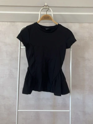 Svart topp med volangdetalj - Snygg svart topp från Gina med korta ärmar och volangdetalj nedtill som ger en feminin touch. Toppen har rund halsringning och är tillverkad i mjukt bomullsmaterial. Perfekt att styla med jeans eller kjol för en trendig look.