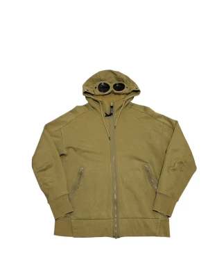 Olivgrön hoodie med glasögon i huvan - Säljer en olivgrön hoodie från C.P. Company med dragkedja framtill och två fickor med dragkedja. Huvan har inbyggda glasögon, vilket ger en unik och cool look. Tillverkad i mjukt bomullsmaterial och har ribbade muddar vid ärmslut och nederkant.                                                                   📍Storlek:M  💪🏽Skick:Mycket bra   📐Mått:Längd:68:cm:Bred:62:cm:  📎Material:100%Bomull:  📦Skickar alltid inom 48h  ✅Äkthetsgaranti