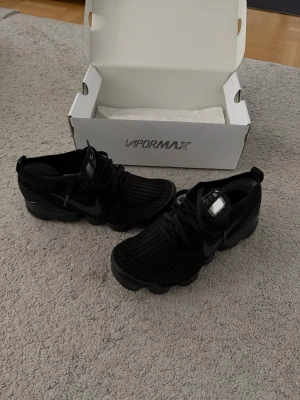 Nike Air Vapormax Flyknit 3 svarta - Svarta Nike Air Vapormax Flyknit sneakers med stickad ovandel och synlig Air-sula. Swoosh-logga på sidan och klassisk Vapormax-design. Skorna har rund tå och svarta skosnören. Perfekta för dig som gillar sportig och modern stil.