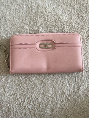 Rosa plånbok från Forever Young - Söt rosa plånbok från Forever Young med silverfärgad metallogo och texten 'Originally from love' framtill. Plånboken har en stilren, rektangulär form och är gjord i ett mjukt, slätt konstläder. Perfekt för dig som gillar gulliga accessoarer och vill ha ordning på kort och kontanter.