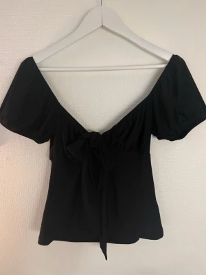 Svart topp med puffärm och rosett - Snygg svart topp med puffiga korta ärmar och en stor rosett framtill. Djup v-ringning och lätt croppad passform. Perfekt för dig som gillar en feminin och trendig stil. Materialet känns mjukt och lätt, troligen polyester.