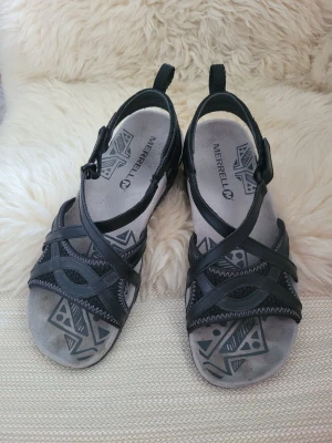 Svarta Merrell sandaler med mönster - Säljer ett par svarta sandaler från Merrell med snygga geometriska mönster på innersulan och robust gummisula med grepp. Sandalerna har flera korsade remmar i syntetmaterial och justerbar rem med spänne runt hälen. Perfekta för sommarens äventyr.