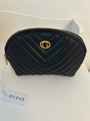 Svart quiltad necessär från Guess - Stilren svart necessär från Guess med quiltad design och guldfärgad dragkedja. Väskan har ett G-emblem i guld på framsidan. Aldrig använd och har prislappen kvar. 