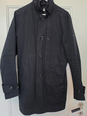 G. Star Raw rock med hög krage - Svart rock med hög krage och knäppning framtill. Rocken har två snedställda fickor och dekorativa knappar på ärmsluten.. Enkel och stilren design som passar till många outfits.