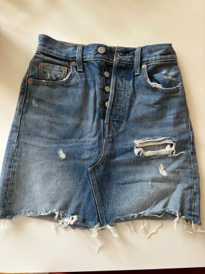 Blå jeanskjol från Levi's med slitningar - Cool kort jeanskjol från Levi's i blå denim med slitna detaljer och fransig nederkant. Kjolen har klassiska fem fickor, knappar framtill och råa kanter för en avslappnad look. 