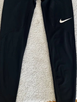 Svarta Nike Dri-Fit tights - Säljer ett par svarta Nike Dri-Fit tights med kompressionspassform. Tightsen har en diskret Nike-logga på benet och bred elastisk midja med grå text. Materialet är polyester och elastan, perfekt för träning och sport.