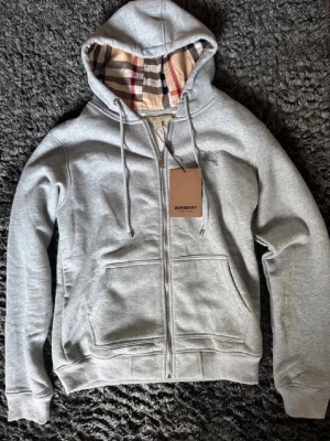 Grå zip hoodie från Burberry - Snygg grå zip hoodie från Burberry med klassiskt rutigt foder i huvan och dragsko. Hoodien har två stora fickor framtill och ribbade muddar vid ärmar och nederkant. Perfekt för en chill och stilren look. Pris kan diskuteras, även öppen för byten, kontakta vid intresse.