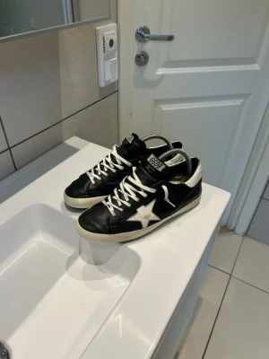 Svarta GGDB sneakers med vit stjärna - Snygga svarta sneakers från Golden Goose med vit stjärndetalj på sidan, vita skosnören och vit sula. Skorna har en klassisk låg profil och GGDB-logga på plösen. Perfekta för dig som gillar streetstyle och vill ha något unikt på fötterna.
