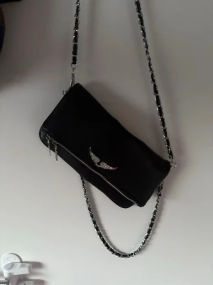 liknande zadig väska - Snygg svart axelväska i konstläder med silverfärgad kedjerem och dragkedja. Väskan har ett emblem med vingar framtill och magnetknappar på insidan. Perfekt för dig som gillar en edgy och stilren look.
