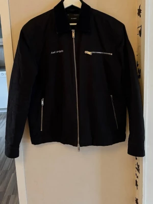 Svart jacka från Axel Arigato - Svart jacka från Axel Arigato med raka linjer, silverfärgade dragkedjor och broderad logga på bröstet. Jackan har klassisk krage i manchester och flera fickor med dragkedja. Perfekt för dig som gillar stilren och modern design. Står XS i den men är som en S för herr. 