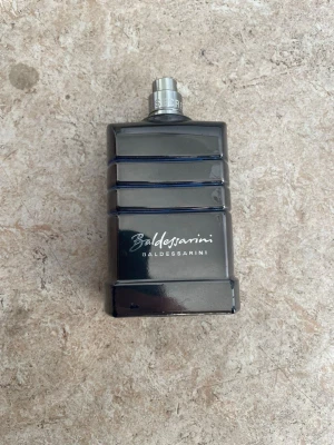 Baldessarini parfym mörkblå flaska - Stilren parfym från Baldessarini i en mörkblå, rektangulär glasflaska med silverfärgad spraykork. Flaskan har horisontella räfflor och loggan tryckt i vitt framtill. Perfekt för dig som gillar exklusiva dofter och snygg design.
