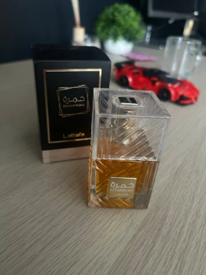 Lattafa Khamrah parfym - Lattafa Khamrah är en lyxig parfym i en elegant, transparent fyrkantig glasflaska med guldtonad vätska. Flaskan har ett geometriskt mönster och en guldig etikett med arabiska och engelska text. Medföljer en svart och guld box som förstärker premiumkänslan.
