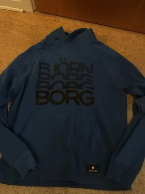 Blå sett från Björn Borg - Snygg blå hoodie från Björn Borg med stort svart tryck på bröstet och klassisk känguruficka. Tröjan har huva och är tillverkad i mjukt material som passar perfekt för en avslappnad stil. Logotypen finns även på en liten patch vid fickan. Och byxor