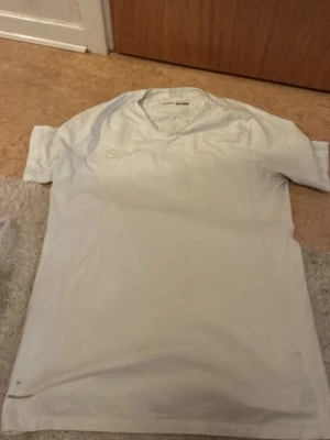 Vit Nike sporttröja med V-ringning - Säljer en vit sporttröja från Nike med V-ringning och korta ärmar. Tröjan har diskret Nike-logga på bröstet och är tillverkad i ett lätt och ventilerande material som passar perfekt för träning och sportaktiviteter.