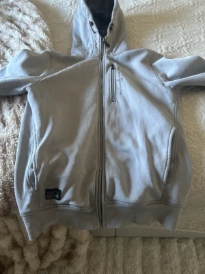 Sail Racing - Ljusgrå hoodie från sail racing med dragkedja framtill och två fickor. Insidan är fodrad med mjuk fleece för extra komfort. Klassisk huva med dragsko och diskret logga på bröstet. Perfekt för chill dagar eller när du vill ha något bekvämt.