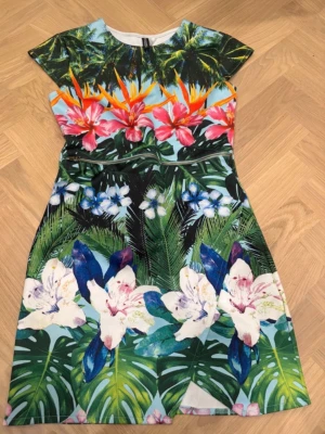 Färgglad blommig kortklänning - Säljer en superfin kortklänning med tropiskt blommönster i grönt, blått, rosa och vitt. Klänningen har rund hals, korta ärmar och är gjord i ett mjukt syntetmaterial. Perfekt för dig som vill sticka ut med färg och mönster!