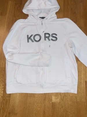 Vit hoodie från Michael Kors - Snygg vit hoodie från Michael Kors med dragkedja och stor broderad KORS-logga i grått på bröstet. Hoodien har huva med dragsko och två fickor framtill. Perfekt för en clean och stilren look. Äkta.