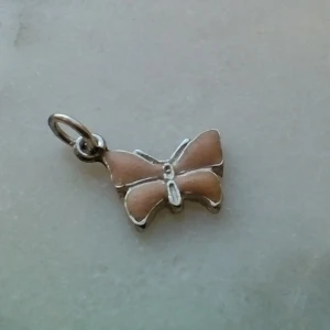 Rosa/periskofärgad liten berlock/hänge - fjäril i silver 925 - Litet hänge i äkta silver. Stämplad 925 och något mer som jag inte kan tyda. Rosa/persikofärgad framsida - ganska matt och lite ojämn yta. Ca 1 cm bred. Frakt 22 kr.