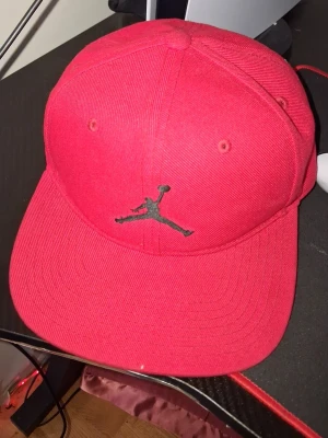 Röd Jordan keps med broderad logga - Säljer en röd keps från Jordan med den klassiska Jumpman-loggan broderad framtill i svart. Kepsen har böjd skärm och ventilationshål. Insidan har texten 'Quality inspired by the greatest player ever'. Tillverkad i 100% polyester.