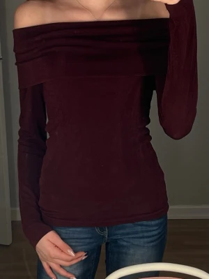 Mörkröd offshoulder topp - Trendig mörkröd offshoulder topp med lång ärm och figurnära passform. Toppen har en bred kant upptill som ger en snygg detalj och är perfekt för dig som vill ha en stilren men ändå iögonfallande look. Materialet känns mjukt och stretchigt💕