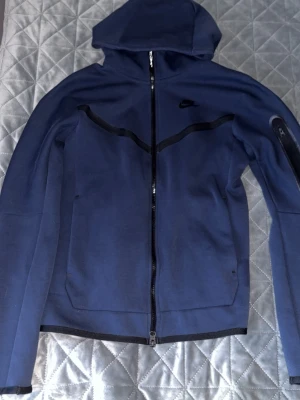 Blå Nike hoodie med dragkedja - Snygg blå hoodie från Nike med hel dragkedja framtill och svart Nike-logga på bröstet. Hoodien har svarta detaljer över bröstet och en ficka med dragkedja på ena ärmen. Tillverkad i ett mjukt och stretchigt material, perfekt för en sportig look.