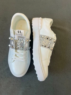 Valentino Rockstud vita sneakers - Snygga vita sneakers från Valentino med breda band över vristen dekorerade med silverfärgade nitar och glitterdetaljer. Skorna har en chunky sula och klassisk låg profil. Perfekta för dig som vill sticka ut med exklusiv design och coola detaljer.