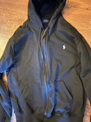 Svart hoodie från Polo Ralph Lauren - Svart hoodie med dragkedja från Polo Ralph Lauren. Klassisk design med vit broderad logga på bröstet. Perfekt för en avslappnad och stilren look. Storlek står som L men passar som M