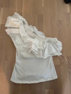 Vit trendig topp med volangdetalj - Trendig vit topp med stora volanger över ena axeln. Asymmetrisk design som ger en cool look. Tillverkad i stretchigt material som sitter snyggt på kroppen. Perfekt att styla med jeans eller kjol för en modern outfit.