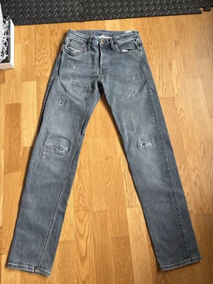 Grå slitna jeans med raka ben - Säljer ett par grå jeans med raka ben och distressed detaljer på knän och lår. Jeansen har klassisk femficksmodell och normal midja. Materialet är denim i bomull och de har en avslappnad, trendig look.