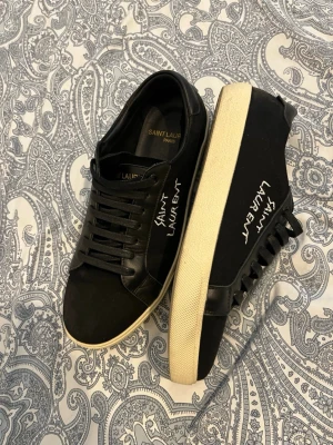 Svarta Saint Laurent sneakers - Snygga svarta sneakers från Saint Laurent med vit broderad logga på sidan. Skorna har svart ovandel i canvas och läder, samt vit sula i gummi. Klassisk låg modell med svarta skosnören och diskret branding på insidan. Perfekta för en stilren och trendig look.