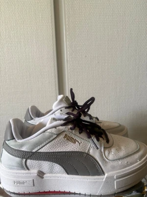 Vita Puma sneakers med grå detaljer - Snygga vita sneakers från Puma med gråa detaljer och svart snörning. Skorna har en chunky sula och klassisk Puma-logga i guld på sidan. Ovandelen är i läder och textil, och designen är lågskuren för en clean och modern look.
