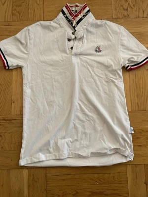 Vit Moncler topp med randig krage - Snygg vit topp från Moncler med korta ärmar och unik randig krage i rött, svart och vitt. Knappdetaljer vid halsen och Moncler-logga på bröstet. Ärmsluten har matchande randiga detaljer. Perfekt för en stilren och sportig look.