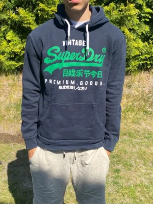 Mörkblå Superdry hoodie med tryck - Snygg mörkblå hoodie från Superdry med stort grönt och vitt tryck på bröstet. Klassisk känguruficka framtill, vita dragsnören i huvan och mjukt material på insidan. Perfekt för dig som gillar streetwear och vill ha en bekväm och stilren look.