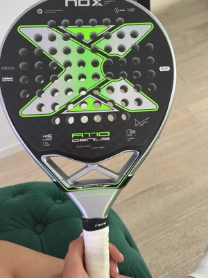 NOX AT10 Genius padelracket - Säljer ett NOX AT10 Genius padelracket med svart och silver design samt gröna detaljer. Racket har en stor X-formad grafik, perforerad träffyta och vitt grepp. Lite färg som gått av på kanten av racket men inga sprickor eller fel på träffytan. Priset är diskuterbart 