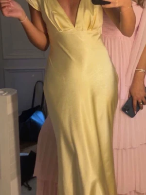 Ljusgul långklänning i satin - Elegant långklänning i ljusgul satin med v-ringning och korta ärmar. Klänningen har en figurnära passform och ett mjukt, glansigt material som ger en lyxig känsla. Perfekt för speciella tillfällen när du vill sticka ut.
