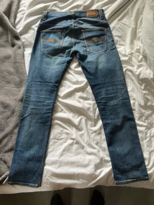 Nudie Jeans - Snygga blå jeans från Nudie Jeans med klassisk femficksdesign och subtila slitningar. Jeansen har kontrastsömmar och läderpatch bak i midjan. Tillverkade i mjuk denim med lite stretch för extra komfort. Modellen är thin finn, men de sitter inte särskilt tight på mig som är 184 och väger 77kg. L32, W31. Jeansen har inga defekter!