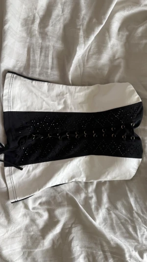 Svartvit korsett med snörning - Säljer en snygg svartvit korsett med snörning framtill och dekorativa små hål i ett diamantmönster. Korsetten har en klassisk form och är tillverkad i bomullsliknande material. Perfekt för att skapa en edgy look.