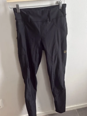 Svarta ridleggings från Maya delorenz - Snygga svarta ridleggings från Maya delorenz med hög midja, bälteshällor och smarta fickor. Stretchigt material som ger bra rörelsefrihet och detaljer med logga på både fram- och baksida. Perfekta för dig som vill ha både stil och funktion i stallet.
